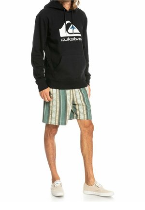 Sort hættetrøje med en hvid og blå Quiksilver-logo, kombineret med grønne og cremefarvede shorts med striber. Beige sneakers fuldender looket.