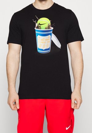 Schwarzes T-Shirt mit Grafik eines blauen Bechers, aus dem Limonade verschüttet wird, und einem neongrünen Nike Tennisball, getragen von einer Person in roten Nike-Shorts.