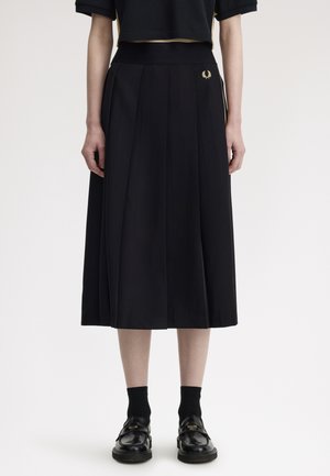 Mujer con falda midi negra plisada con un pequeño logo dorado, top corto negro, calcetines negros y mocasines negros sobre un fondo blanco.