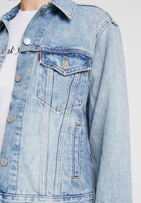 Světlá modrá denimová bunda s klasickým límcem, kovovými knoflíky, dvěma náprsními kapsami a jemnou vybledlou strukturou. Výrazná červená nášivka na kapse.