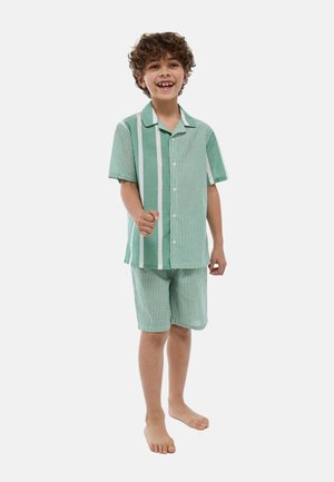 Garçon souriant pieds nus debout, portant une chemise à rayures vertes et blanches à manches courtes boutonnée et un short assorti.