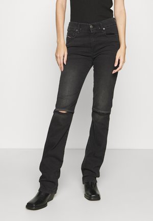 Personne portant un jean noir avec des découpes aux genoux, des bottines noires et un haut sans manches noir, debout sur un fond blanc uni.