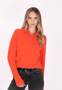Langarmige, rits-top in feloranje materiaal, met een aansluitende pasvorm. Gepaard met zwarte jeans en een ronde gespsluiting.