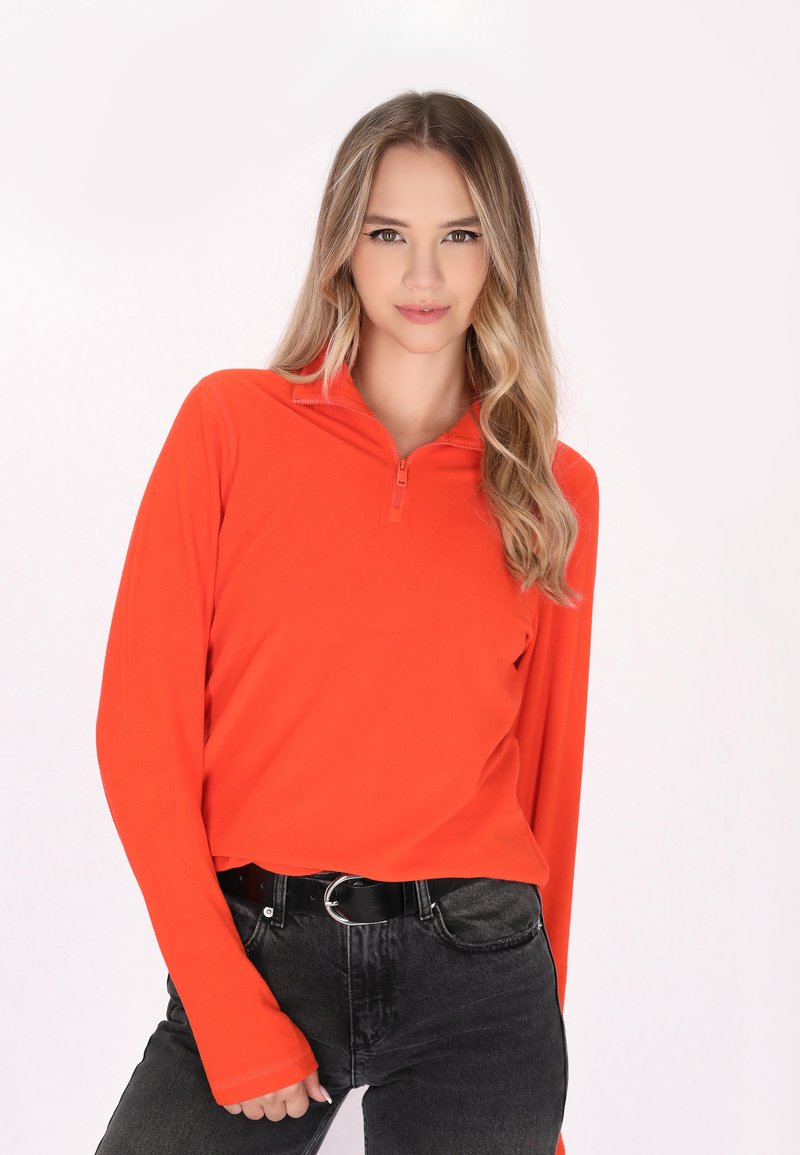 Langarmige, rits-top in feloranje materiaal, met een aansluitende pasvorm. Gepaard met zwarte jeans en een ronde gespsluiting.