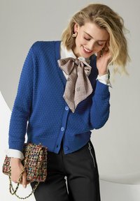 Vrouw met blond haar die een gestructureerde blauwe cardigan, een witte blouse met een patroonstrik, zwarte broek draagt en een kleurrijke gevlochten handtas vasthoudt.