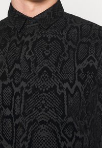 Camicia nera caratterizzata da un sottile motivo testurizzato a pelle di serpente. Presenta un colletto a punta e un unico bottone nero al collo.