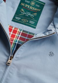 Lyseblå jakke med bronze lynlås delvist åben, der viser rød og blå tartan-foring samt grøn Superdry mærkat inde i kraven.