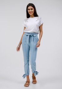 T-shirt bianco con ricami floreali sulle spalle, abbinato a pantaloni azzurri chiari a vita alta con cintura annodata e dettagli a fiocco sulle caviglie.
