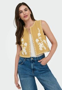 Gul kort ärm med vit blommig broderi, scallopade kanter och öppen framsida, lager över en vit topp och tillsammans med blå jeans.