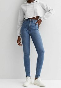 Istuvien sinisten farkkujen, joissa on korkeavyötärö, hienovarainen kulutus ja klassinen viiden taskun malli, yhdistettynä lyhennettyyn harmaaseen huppariin ja valkoisiin tennareihin.