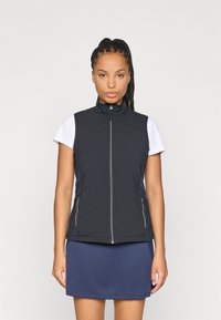 Sort quiltet vest med høy krage, med glidelåslommer og en tettsittende design, båret over en hvit T-skjorte med korte ermer og et blårutete skjørt.