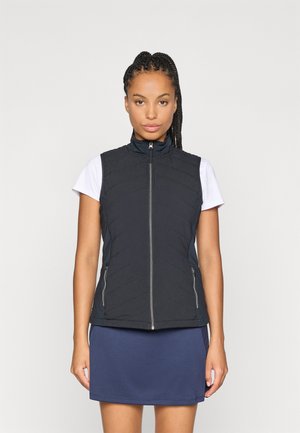 Black quilted vest with high collar, featuring zippered pockets and a fitted design, worn over a white short-sleeve top and blue skirt.