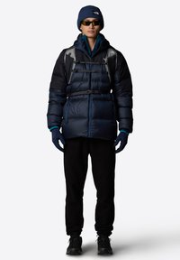 Granatowa kurtka puchowa z poziomym wzorem na wypustkach, czarne spodnie z polaru, niebieskie rękawice i czapka; uzupełnione o okulary przeciwsłoneczne i plecak.