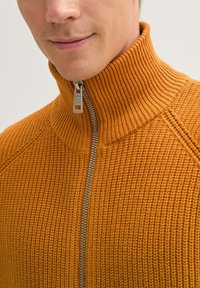 Oranger Strick-Zip-Sweater mit hohem Kragen und strukturiertem Stoff; verfügt über einen silbernen Reißverschluss und gerippte Akzente am Kragen.