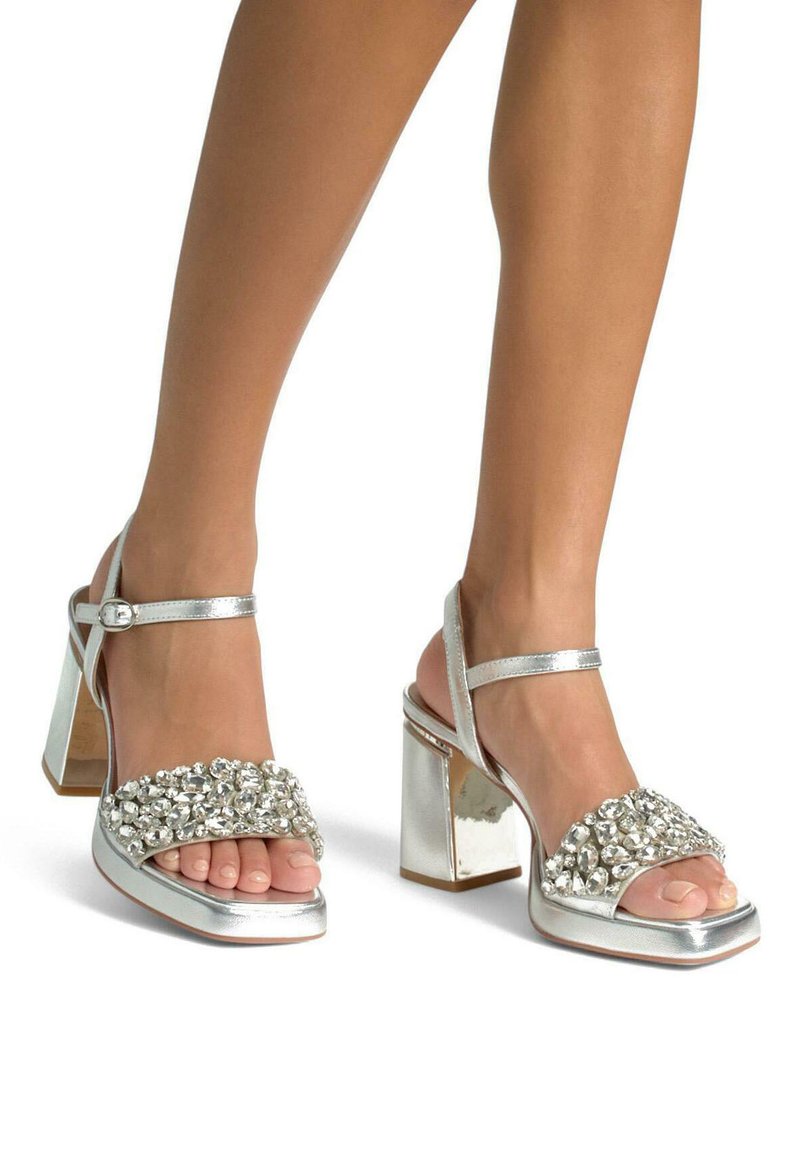 Sandali alti argentati con tacco a blocco spesso, caratterizzati da un ampia fascia decorata con gemme trasparenti e una finitura metallica.