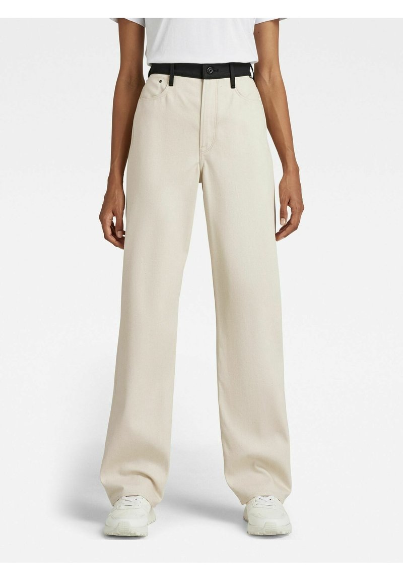 Pantalones de pierna ancha beige con un cinturón negro, que presentan un cierre de botón y cremallera, bolsillos frontales y una textura suave. Combinados con zapatillas blancas.