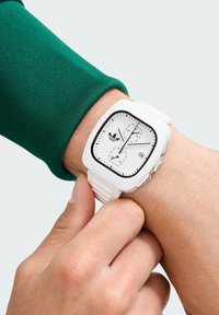 Ceas alb pătrat cu accente negre, curea din silicoan texturată și cadran alb cu trei subdialuri și o fereastră de dată, purtat pe un încheietură.