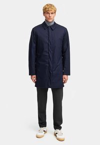 Marinblå lång kappa i slät tyg med knäppning framtill, krage och långa ärmar, kombinerad med mörka byxor och sneakers.