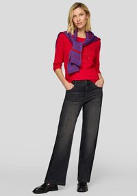 Rotes Langarmhemd mit Logodetail, kombiniert mit weiten schwarzen Jeans. Ein lila karierter Schal liegt über den Schultern. Schwarze Schuhe runden das Outfit ab.