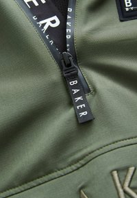 Top in tessuto verde oliva con zip, dotato di un'etichetta nera con "BAKER." Texture liscia e dettagli cuciti nell'area della zip.