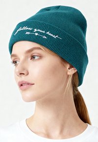 Cappello a cuffia in maglia costina color verde smeraldo con testo ricamato bianco "segui il tuo cuore" e design a freccia. Stile aderente, texture liscia e materiale morbido.