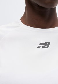 Κοντινό πλάνο ενός ατόμου που φοράει ένα λευκό αθλητικό μπλουζάκι με το μαύρο λογότυπο της New Balance στο επάνω μέρος του στήθους.