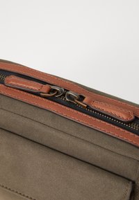 Sac en toile avec un accent en cuir marron au niveau de la fermeture éclair, une fermeture à glissière et une texture lisse avec de subtils points de couture diagonaux.