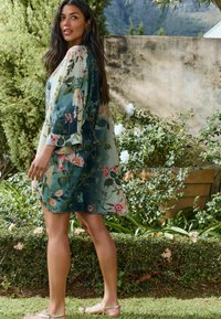 A woman with long hair stands outdoors, wearing a floral wrap dress, next to greenery and blooming flowers in a garden setting.