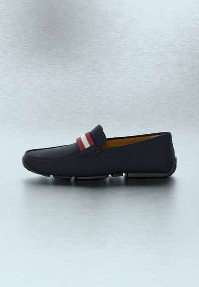Mocassin en cuir noir avec strap rayé rouge et blanc, couture mocassin, et semelle en caoutchouc, présenté sur une surface texturée gris clair.