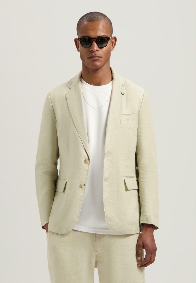 Lichtbeige blazer van linnen met twee voorkruiken, met een notch revers en een knoopsluiting. Draagt over een wit shirt en beige broek.
