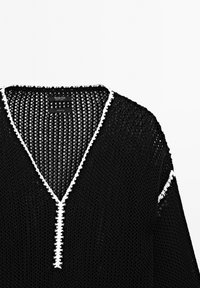Pull en maille ouverte noir avec décolleté en V et bord blanc le long de l'encolure et des manches, montrant l'étiquette de la marque à l'intérieur du col.