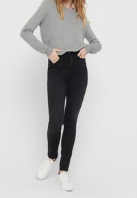 Schwarze, hoch taillierte Jeans aus Denim mit schmaler Passform und minimalen Details, kombiniert mit einem hellgrauen Strickpullover und weißen Sneakern.