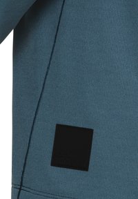 Tessuto blu testurizzato con un sottile motivo a coste, caratterizzato da una toppa rettangolare nera decorata con il logo in rilievo "Jack Wolfskin" nella parte inferiore.