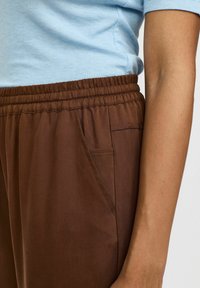 Bruine, lichte, elastische broek met zijzakdetails, gecombineerd met een lichtblauw shirt met korte mouwen, dat een gladde textuur toont.