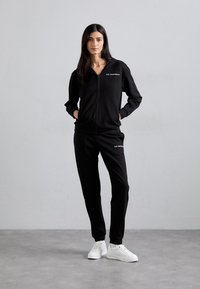Zwarte zip-up sweatshirt met bijpassende broek, beide gemaakt van gladde stof, met een capuchon en logo op de borst, gecombineerd met witte sneakers.