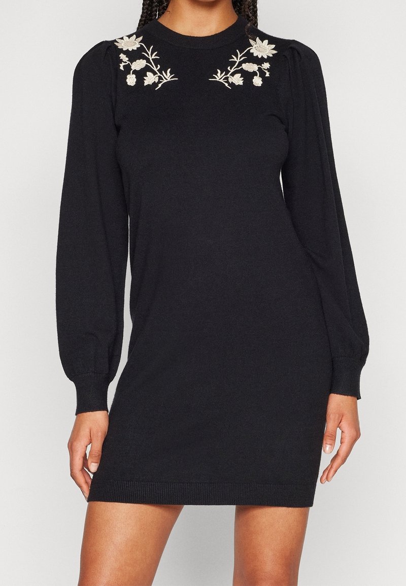Mini-robe noire à manches longues avec broderie florale blanche près de l'encolure, portée par une personne debout avec les mains détendues le long du corps.