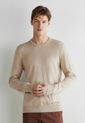 Ung mand stående mod hvid væg, iført beige langærmet sweater og brune bukser, hænder foldet ved taljen, neutral udtryk.