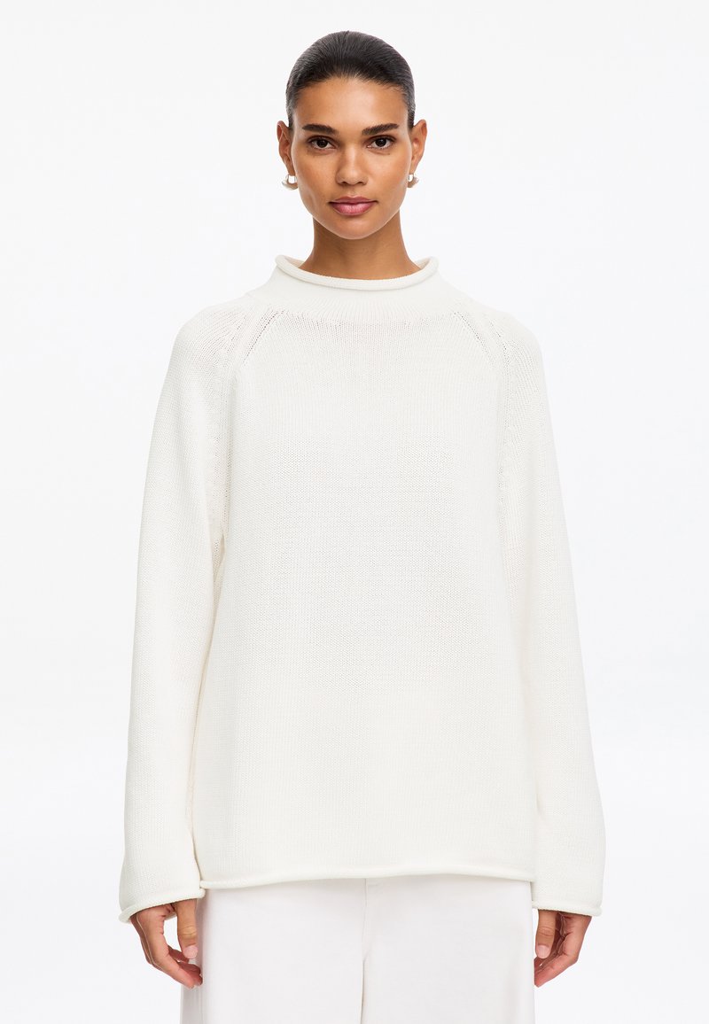 Femme aux cheveux foncés portant un pull en maille blanc et un pantalon blanc, debout devant un fond blanc uni.