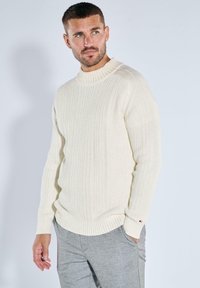 Cremefarbener gerippter Strickpullover mit hohem Kragen und langen Ärmeln, der aus einem strukturierten Stoff gefertigt ist und ein kleines Logo-Akzent nahe dem Bund aufweist.