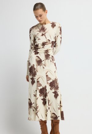 Lange mouw bloemendress in crème met een bruine print. Heeft omgeplooide details in de taille en een vloeiende rok. Gladde stof en getailleerd ontwerp.