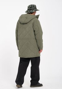 Olijfgroene gewatteerde jas met capuchon, met een boxy pasvorm en een gestructureerd patroon. Combinatie met zwarte wijde broek en een gevlekt emmerhoed.