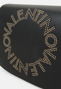 Svart läderhandväska med guldfärgade nitar som bildar ett cirkulärt mönster. Texturerad yta med släta kanter och minimal detaljering.