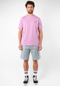 T-shirt en coton violet clair avec un petit motif floral. Short de sport gris avec un logo sur le côté, associé à des baskets noires et des chaussettes à motifs.