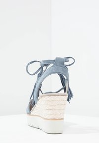 Wedge sandal in soft blue suede with adjustable laces, featuring a woven straw platform and textured beige sole, highlighting a layered design.