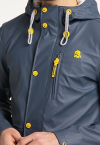 Veste imperméable bleue marine avec un logo jaune, des boutons jaunes contrastants, du matériel en argent et une capuche à cordon de serrage. Poche zippée sur la poitrine.