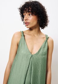 Helles grünes ärmelloses Kleid mit V-Ausschnitt, strukturiertem Stoff, Knotenbändern und plissierten Details an der Vorderseite.