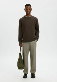 Maglione lavorato a maglia marrone con orlo a coste, sottoshirt grigio chiaro, pantaloni beige e borsa in tessuto verde. Scarpe nere completano l'outfit.