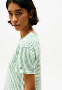 Camiseta de manga corta a rayas en blanco y verde, con un cuello redondo y un pequeño logotipo en la manga. Material de algodón suave.