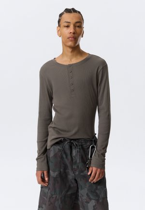 Jeune personne aux cheveux tressés portant une chemise henley à manches longues gris foncé et un pantalon cargo camouflage avec des clés attachées sur le côté.