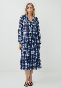 Blusa blu tie-dye con colletto arricciato e maniche lunghe, abbinata a una gonna coordinata. Realizzata in un tessuto leggero e fluido. Sandali neri indossati.